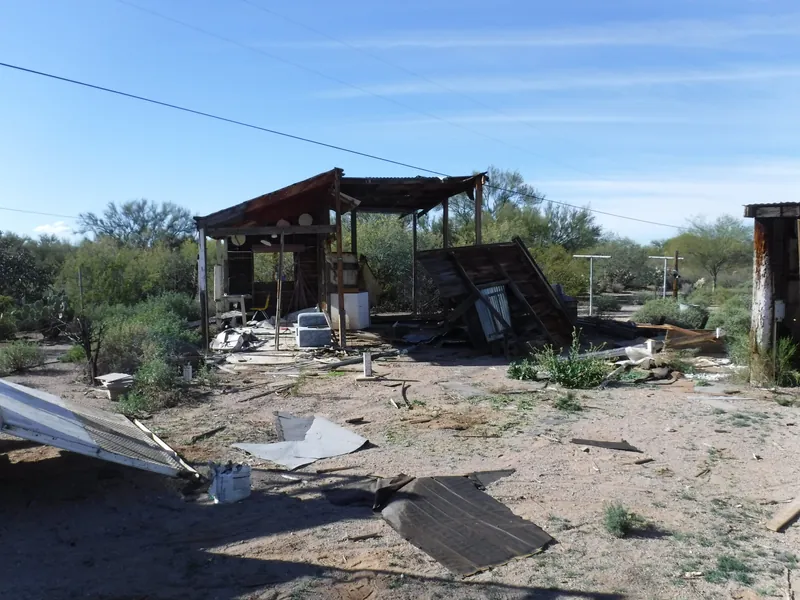 Storm cleanup, before: a storm damaged shed with a collapsed wall and roofing, tar paper and lumber scattered across a desert yard